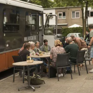Foto van een mobiele ontmoetingsplek. Mensen zitten aan tafeltjes bij een busje.